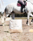 Jutetasche mit schwarzem Motiv auf einem Reitplatz vor einem weißen Pferd