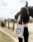 Junge Frau mit heller Baumwolltasche neben einem braunen Pferd auf einem Feldweg