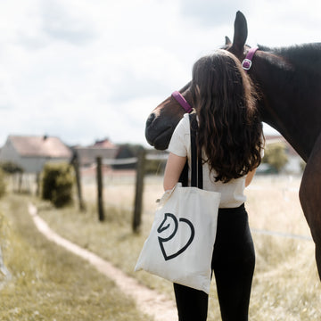 Junge Frau mit heller Baumwolltasche neben einem braunen Pferd auf einem Feldweg